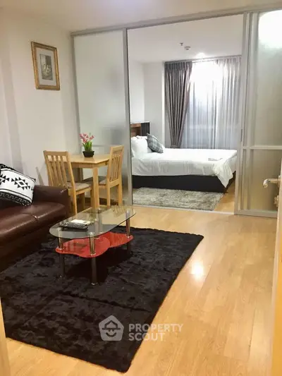 Cozy living room with wooden flooring and glass coffee table leading to a stylish bedroom with plush bedding.