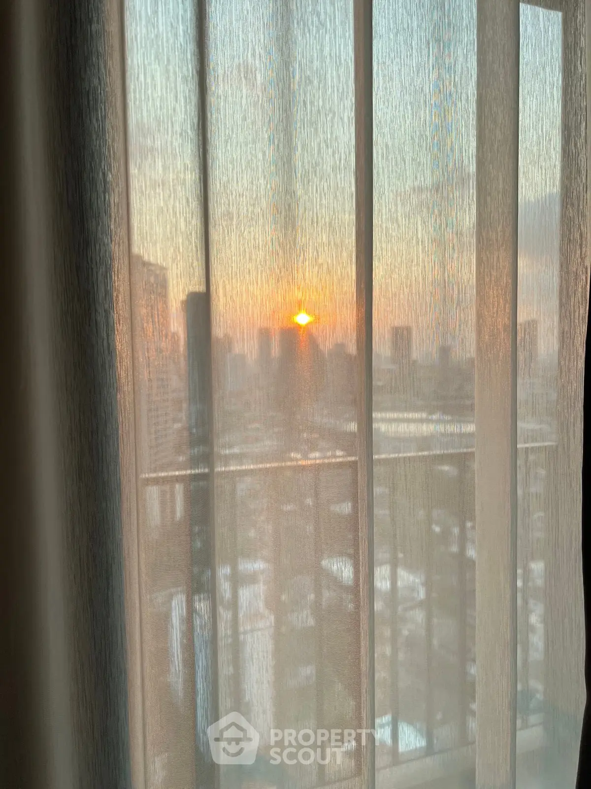 Stunning city view from a high-rise apartment with sunset through sheer curtains.