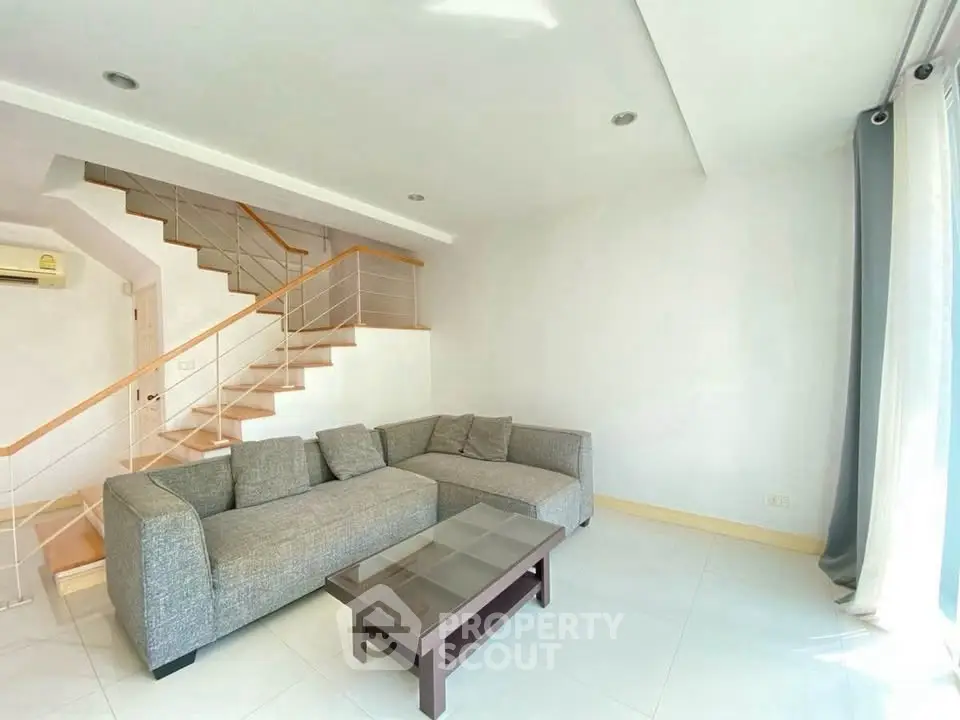 Spacious living room with modern gray sofa and glass coffee table, featuring a stylish staircase and natural light.