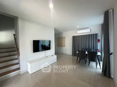 Modern living room with sleek furniture and natural light, featuring a wall-mounted TV and dining area.