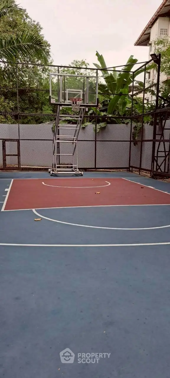 Outdoor basketball court in a residential complex with lush greenery and secure fencing.