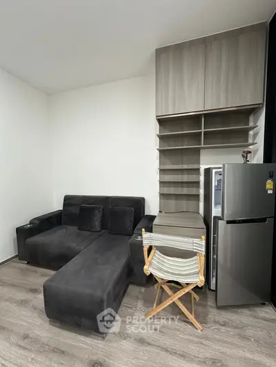Modern living room with sleek black sofa and compact kitchen area featuring a fridge and shelving.