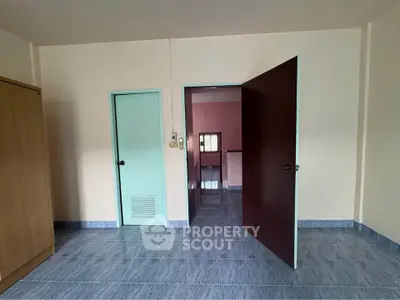 Spacious room with blue tiled flooring and wooden wardrobe, featuring open doors leading to hallway.