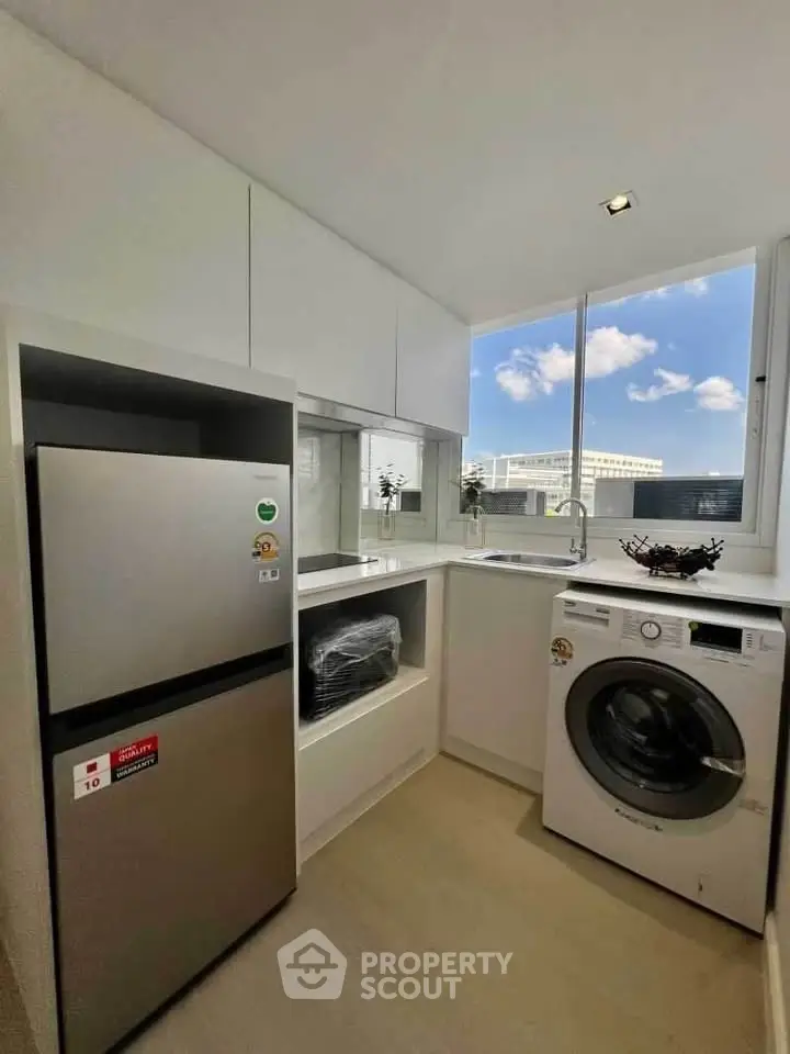 Modern kitchen with washing machine and fridge, bright natural light, sleek white cabinetry.