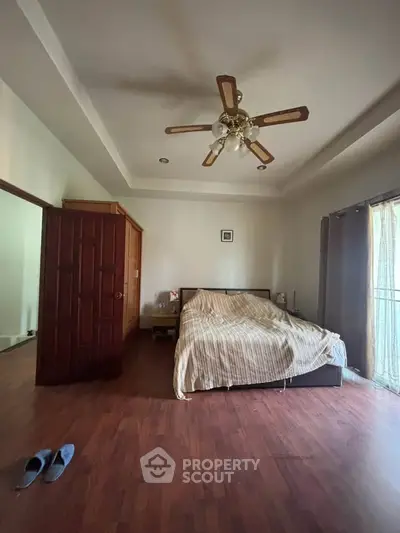 Spacious bedroom with wooden flooring and ceiling fan, featuring natural light from large window.