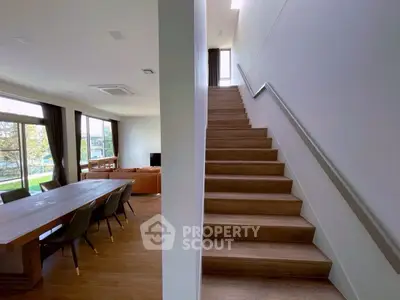 Spacious modern living room with dining area and staircase, featuring large windows and wooden flooring.