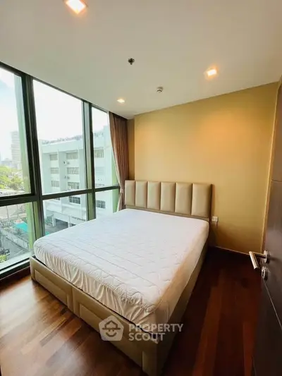 Modern bedroom with large windows and city view, featuring a stylish bed and wooden flooring.