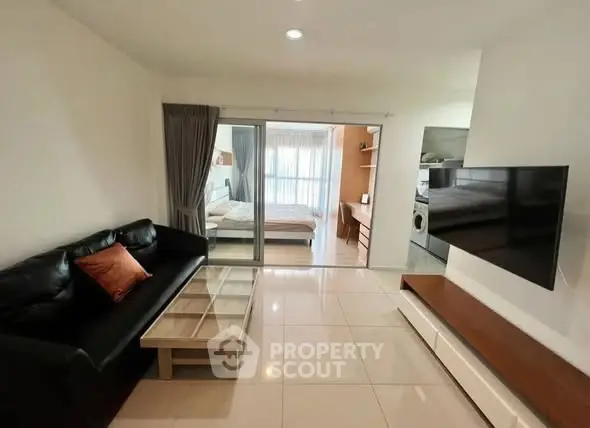 Modern living room with sleek black sofa, glass coffee table, and wall-mounted TV, leading to a cozy bedroom.