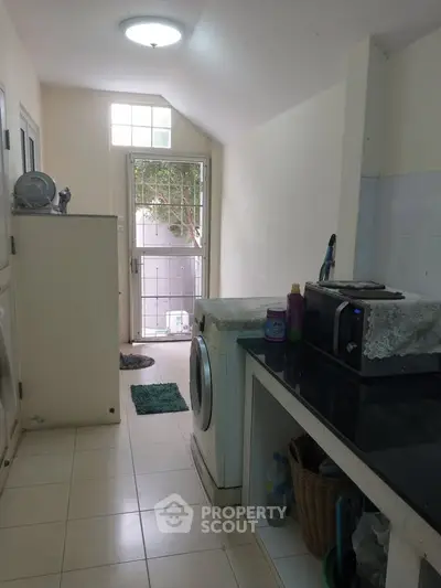 Compact kitchen with washing machine and microwave, bright natural light from window, tiled flooring.