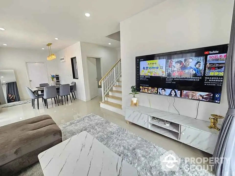 Modern living room with stylish furniture and large TV, open dining area, and staircase.