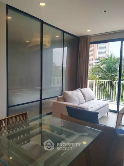 Modern living room with glass table and cozy sofa, featuring a balcony view of lush greenery and cityscape.