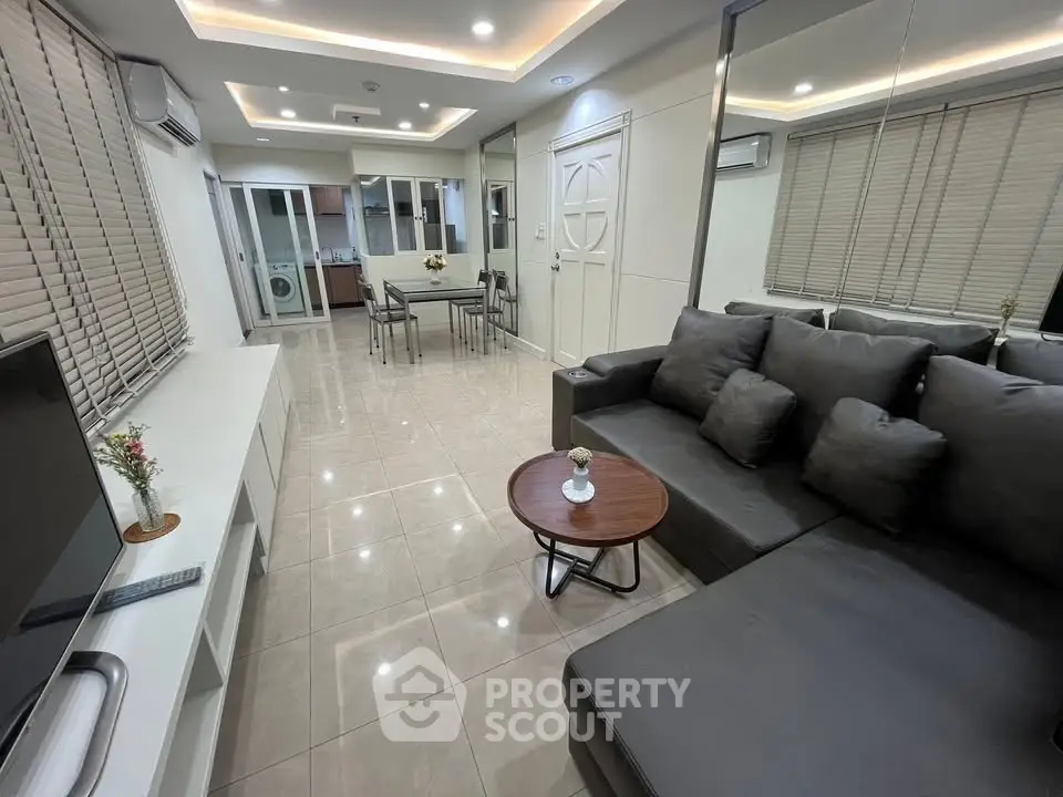 Modern living room with sleek gray sofa and open layout, featuring a dining area and washing machine in the background.