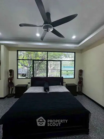 Spacious bedroom with large window and modern ceiling fan, offering a serene view of lush greenery.