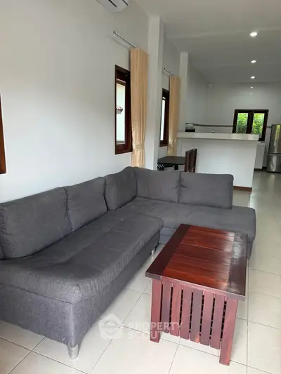 Spacious living room with modern gray sofa and wooden coffee table, open layout to kitchen area.