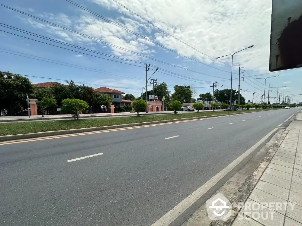 Spacious road view with residential properties and lush greenery under a clear blue sky.