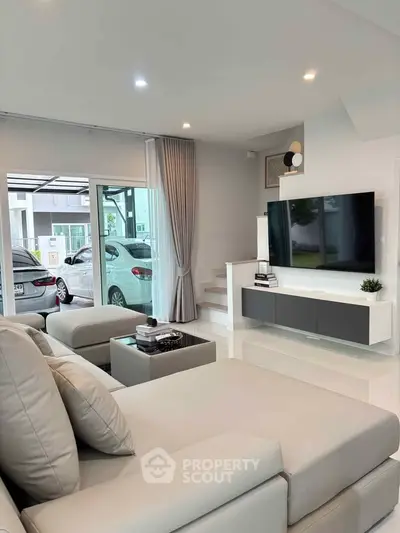 Modern living room with sleek furniture and large TV, featuring a view of the carport through glass doors.