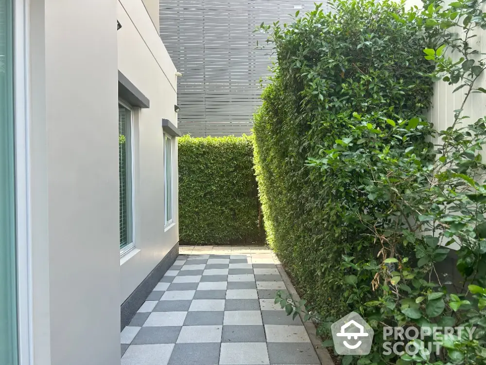 Charming private patio with checkered flooring and lush green hedges, offering a serene outdoor retreat adjacent to a modern home.