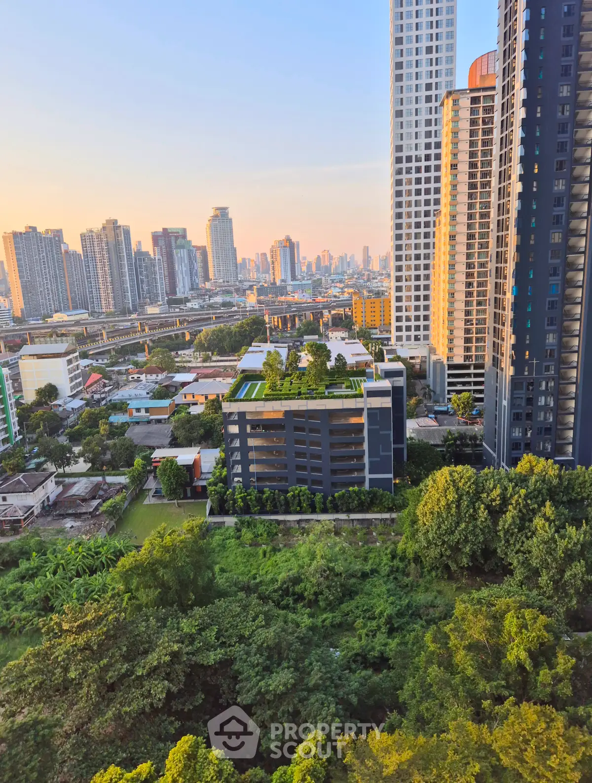 Stunning cityscape view from high-rise apartment with lush greenery and modern buildings at sunset.