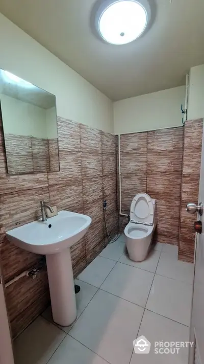 Compact bathroom with modern tiled walls, pedestal sink, and well-maintained toilet under bright ceiling light.