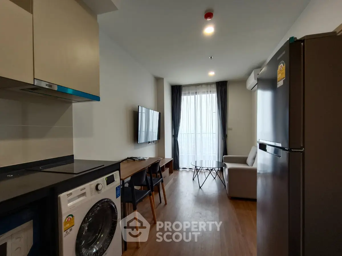 Modern apartment living room with kitchen and washing machine, featuring sleek furniture and natural light.