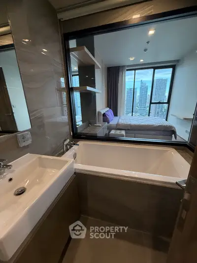 Modern bathroom with bathtub and city view through window into bedroom.