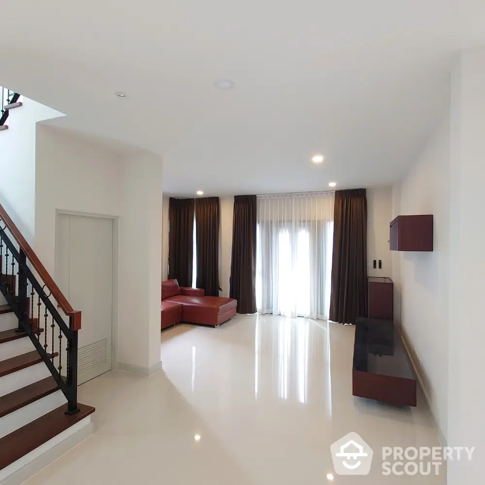 Spacious living room with gleaming white tiles, elegant dark curtains, and a striking red leather sofa set, complemented by a sleek staircase and modern furnishings.