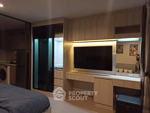 Modern studio apartment with sleek wooden cabinetry and built-in TV, featuring a cozy bed and compact kitchen area.