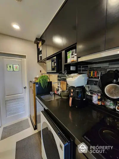 Modern kitchen with sleek black cabinets and appliances, featuring a washing machine and coffee maker.