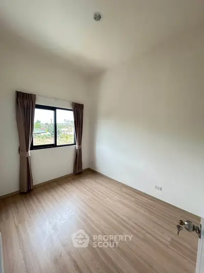 Bright empty room with wooden flooring and large window offering scenic view.