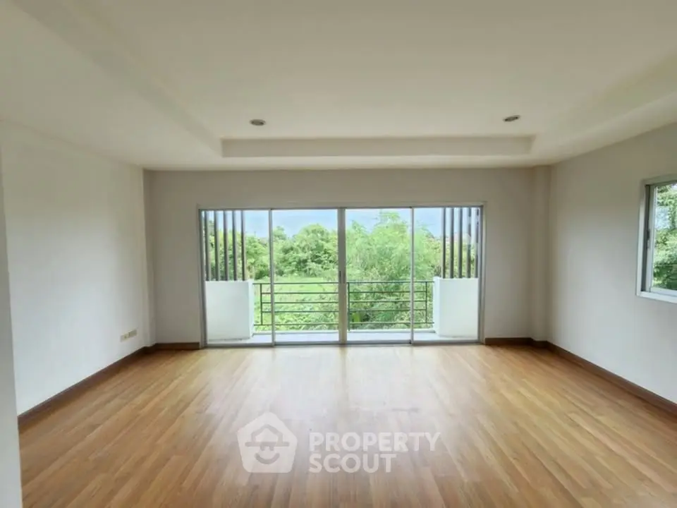 Spacious empty room with wooden flooring and large windows offering lush green views.