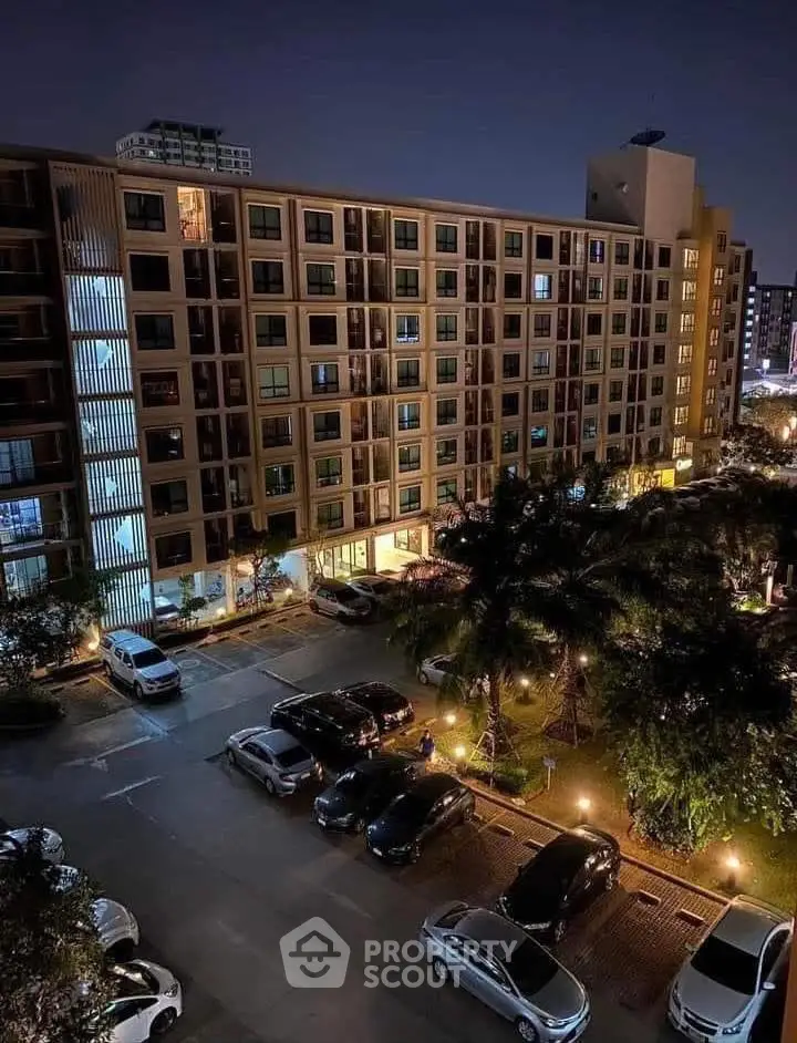 Night view of modern apartment building with well-lit parking area and landscaped surroundings.