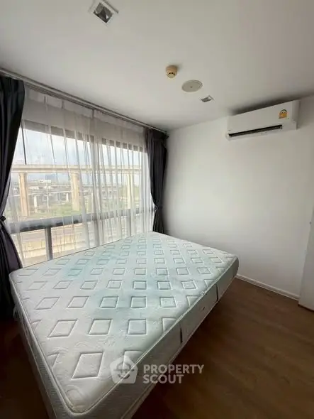 Bright bedroom with large window and air conditioning, featuring a comfortable mattress and wooden flooring.