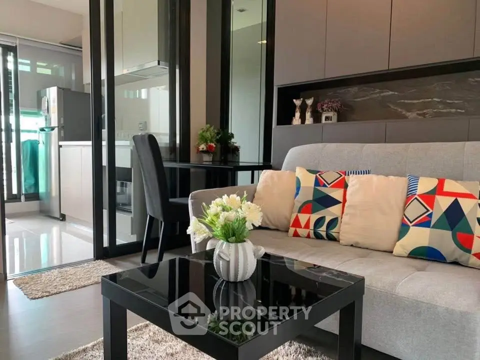 Modern living room with stylish decor and open kitchen layout, featuring colorful cushions and a sleek glass table.
