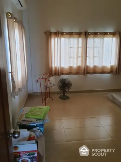 Spacious and well-lit living room with large windows draped in golden curtains, tiled flooring, and a vintage standing fan, awaiting personal touches.