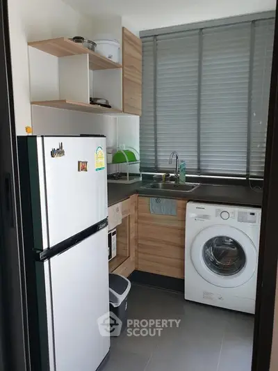 Modern kitchen with washing machine and fridge, featuring sleek wooden cabinets and large window blinds.