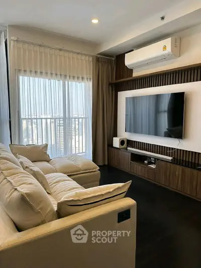 Modern living room with large TV, cozy sofa, and natural light from floor-to-ceiling windows.
