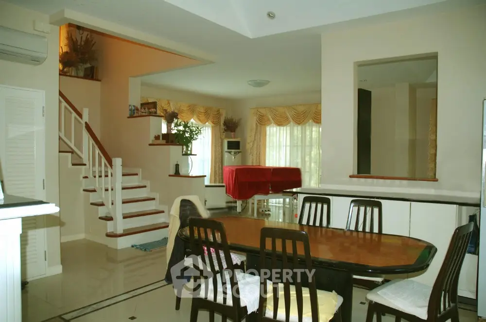Spacious dining area with elegant wooden table set, adjacent to a cozy living space with a grand staircase, bathed in natural light from large windows.