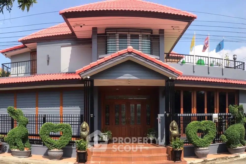Charming two-story house with red-tiled roof and manicured garden, featuring heart-shaped topiary and elegant entrance.