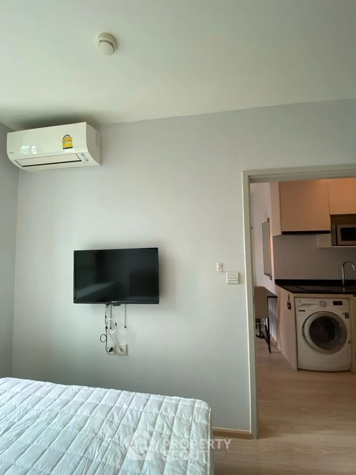 Modern bedroom with wall-mounted TV and view into kitchen with washing machine.