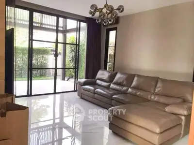 Spacious living room with large leather sectional and floor-to-ceiling windows opening to a lush garden view.