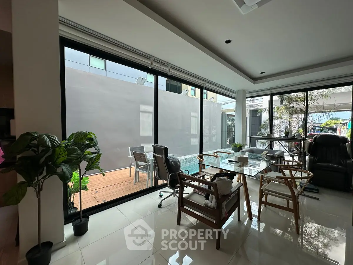 Modern living room with glass walls and outdoor deck, featuring stylish furniture and lush plants.