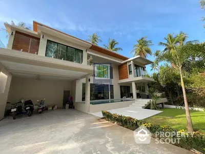 Luxurious modern villa with expansive glass windows, lush greenery, and a spacious driveway, set under a clear blue sky.