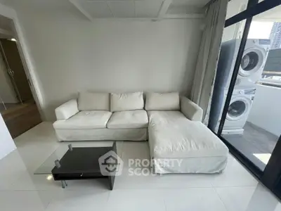Modern living room with white sectional sofa and glass coffee table, featuring a balcony with a stacked washer and dryer.