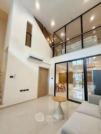 Modern living room with high ceiling and glass railing, open to dining area.