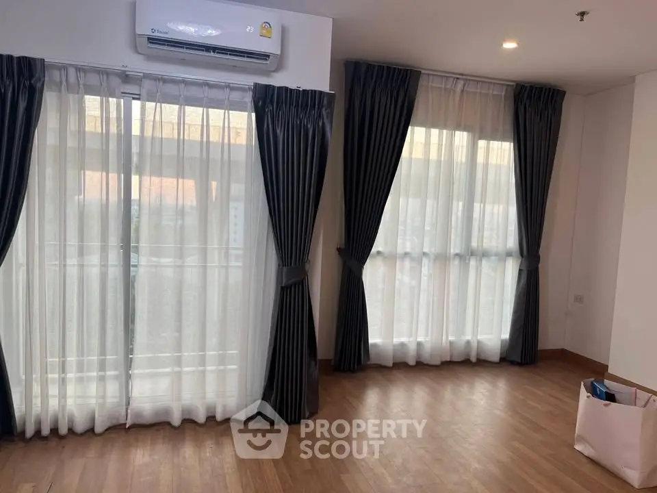 Spacious living room with large windows and elegant curtains, featuring hardwood floors and air conditioning.