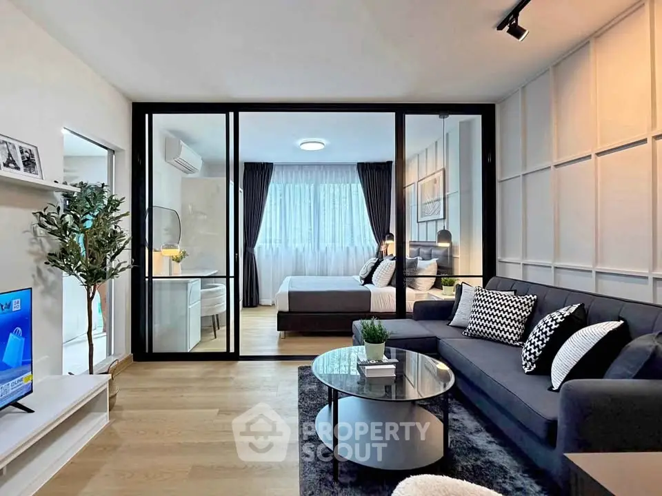Modern open-plan living room with glass partition leading to a stylish bedroom, featuring sleek decor and natural light.