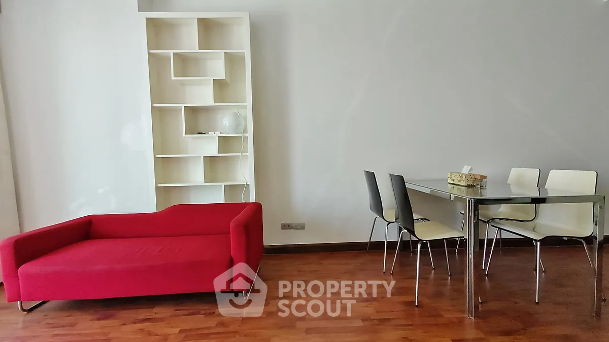 Modern living room with red sofa, glass dining table, and sleek shelving unit on wooden flooring.