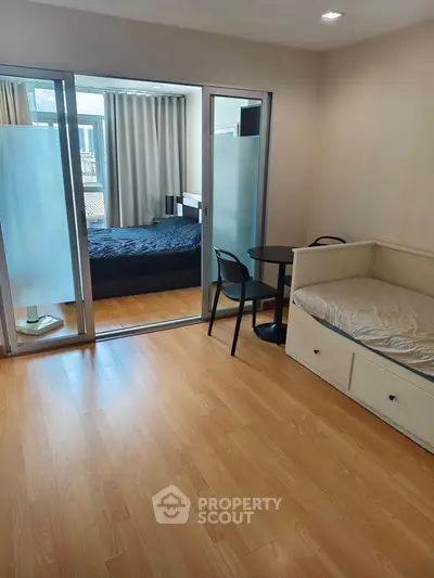 Modern studio apartment with wooden flooring and glass partition, featuring a cozy bed and seating area.