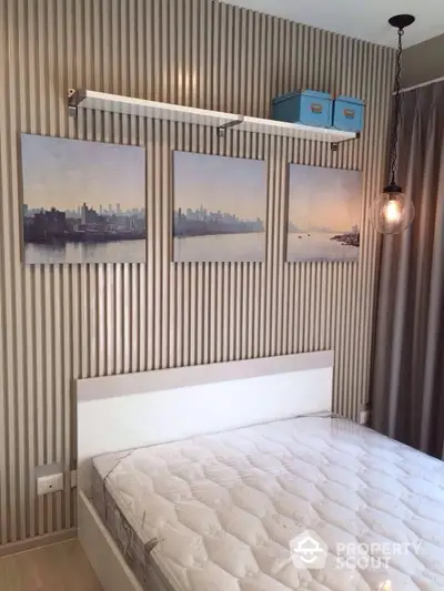 Cozy bedroom with stylish striped wallpaper and cityscape triptych art, complemented by a unique hanging light bulb and a vintage suitcase decor on the upper shelf.