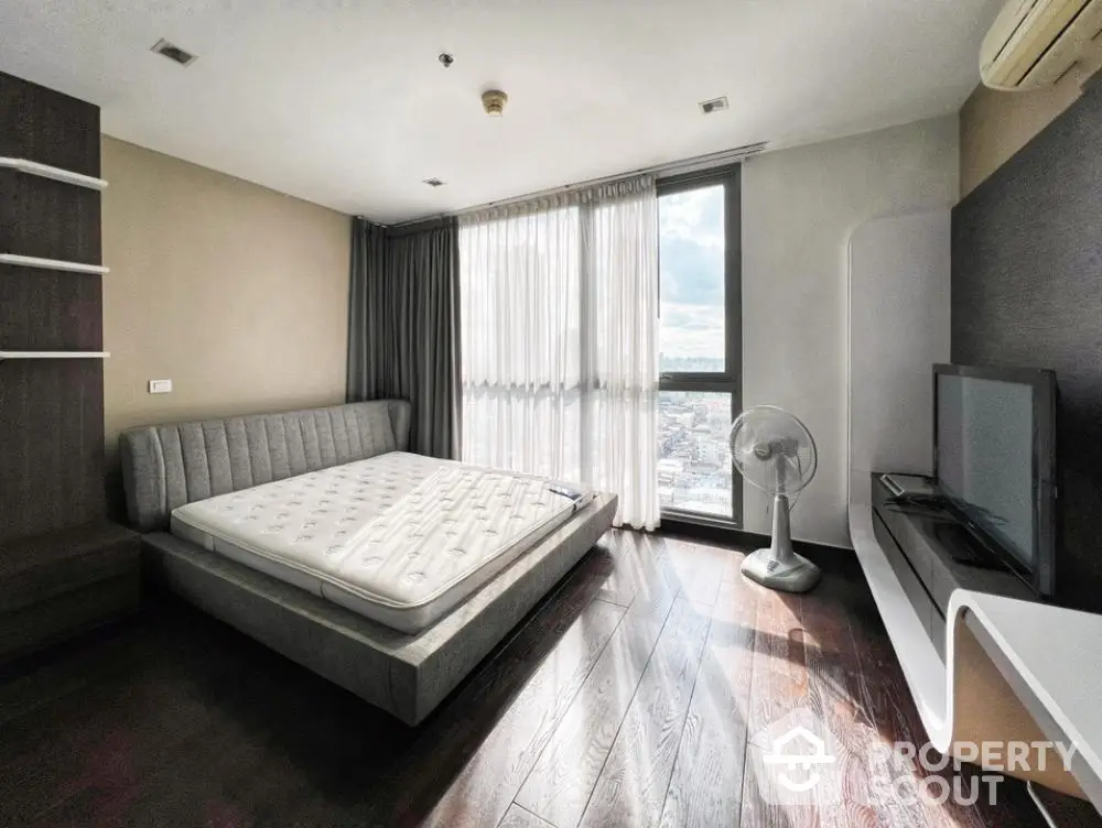 Modern bedroom with large window and city view, featuring sleek furniture and wooden flooring.